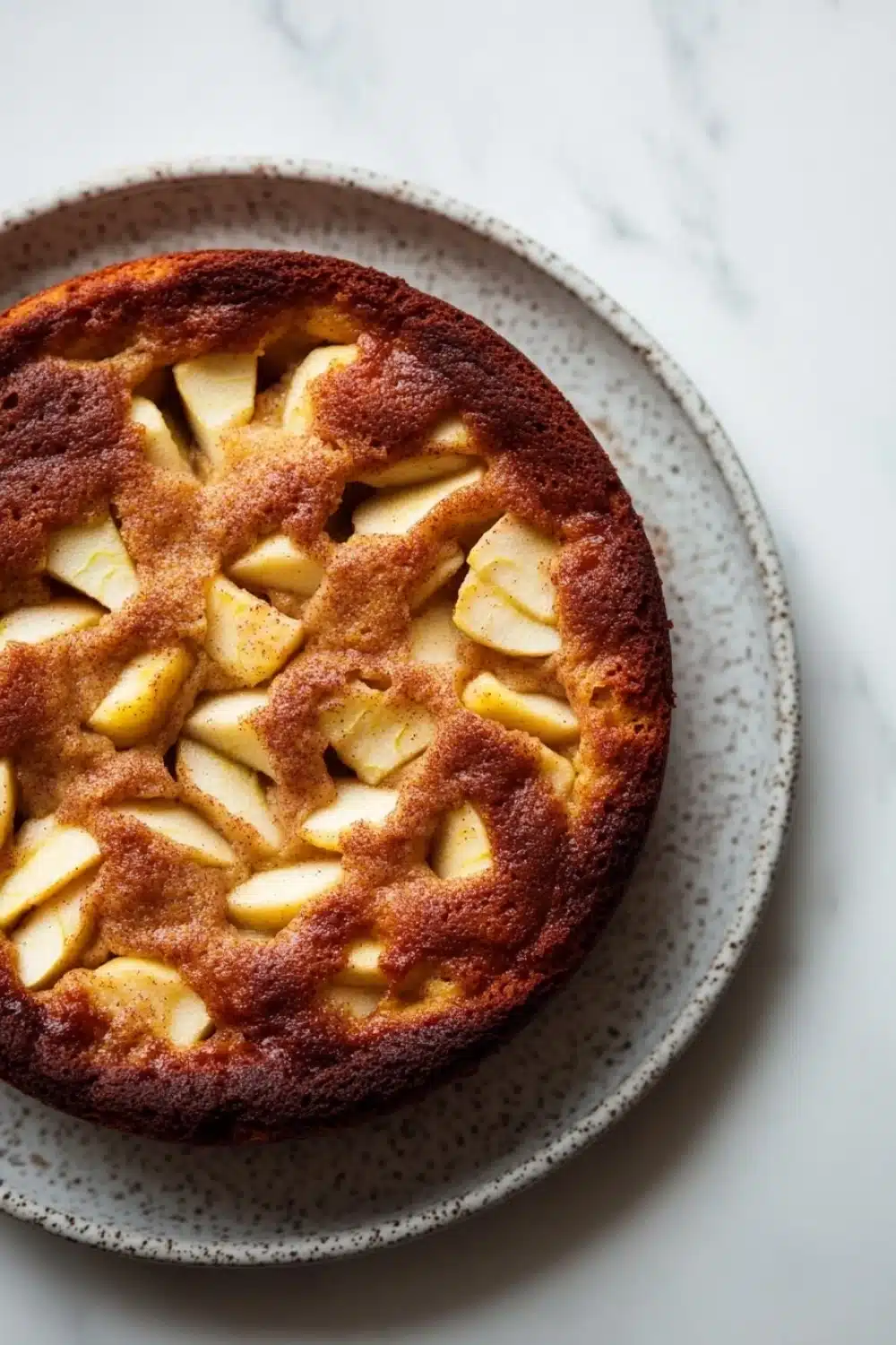 Apple cake no milk - the image is a close-up of a round cake on a white marble countertop. the cake appears to be freshly baked and has a golden brown crust on top. it is topped with sliced apples arranged in a criss-cross pattern, creating a lattice-like design. the apples are cut into small cubes and are arranged evenly across the top of the cake. the plate is round and has small speckles around the edges. the background is blurred, making the cake the focal point of the image.