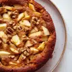Apple cake no milk - the image is a close-up of a round cake on a white marble countertop. the cake appears to be freshly baked and has a golden brown crust on top. it is topped with sliced apples and walnuts, giving it a crunchy texture. the apples are arranged in a circular pattern, with some overlapping each other. the walnuts are scattered throughout the cake, adding a pop of color and texture to the dish. the plate it is sitting on is round and has an orange rim.