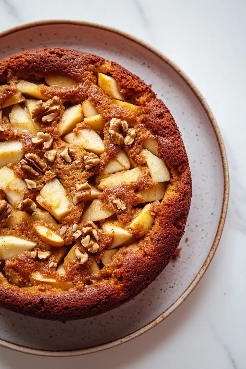 Apple cake no milk - the image is a close-up of a round cake on a white marble countertop. the cake appears to be freshly baked and has a golden brown crust on top. it is topped with sliced apples and walnuts, giving it a crunchy texture. the apples are arranged in a circular pattern, with some overlapping each other. the walnuts are scattered throughout the cake, adding a pop of color and texture to the dish. the plate it is sitting on is round and has an orange rim.