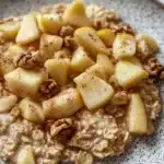 Apple cake oats - the image is a close-up of a bowl of oatmeal with sliced apples and walnuts on top. the oatmeal is a light brown color and appears to be thick and fluffy. the apples are cut into small cubes and are arranged in a circular pattern on top of the oatmeal. the walnuts are scattered throughout the dish, adding a crunchy texture and nutty flavor. the bowl is white with a speckled pattern around the edges. the background is blurred, making the bowl the focal point of the image.