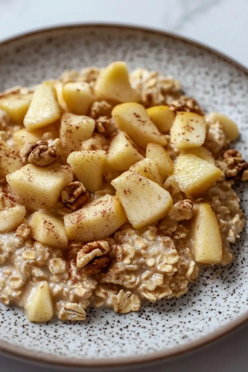 Apple cake oats - the image is a close-up of a bowl of oatmeal with sliced apples and walnuts on top. the oatmeal is a light brown color and appears to be thick and fluffy. the apples are cut into small cubes and are arranged in a circular pattern on top of the oatmeal. the walnuts are scattered throughout the dish, adding a crunchy texture and nutty flavor. the bowl is white with a speckled pattern around the edges. the background is blurred, making the bowl the focal point of the image.
