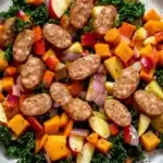 Apple chicken sausage and harvest vegetable skillet - the image is a close-up of a plate of food. the plate is white and round, and it is placed on a wooden table. the food appears to be a salad with a variety of ingredients, including kale, diced carrots, apples, and red onions. there are also small pieces of sausage scattered throughout the salad. the colors of the ingredients are vibrant and the dish looks fresh and appetizing.