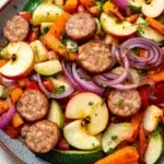 Apple chicken sausage and harvest vegetable skillet - the image is a close-up of a colorful salad in a bowl. the salad is made up of various vegetables, including sliced apples, red onions, carrots, zucchini, and bell peppers. the vegetables are arranged in a circular pattern, with some overlapping each other. the colors of the vegetables are vibrant and the dish looks fresh and appetizing. the bowl is sitting on a wooden table, and the background is blurred, making the salad the focal point of the image.