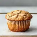 Apple muffin with almond flour - the image shows a freshly baked muffin on a white marble countertop. the muffin is golden brown in color and has a crumbly texture. it is topped with sliced almonds, giving it a crunchy appearance. the edges of the muffin are lined with white paper liners. the background is blurred, making the muffins the focal point of the image.