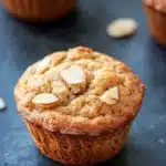 Apple muffin with almond flour - the image shows three freshly baked muffins on a black slate surface. the muffins are golden brown in color and have a crumbly texture. they are topped with sliced almonds, giving them a crunchy appearance. the background is blurred, but it appears to be a kitchen countertop with more muffins scattered around.