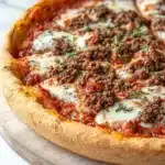 At home deep dish pizza - the image is of a freshly baked pizza on a wooden cutting board. the pizza has a golden brown crust and is topped with melted cheese and chunks of ground meat. the cheese is melted and bubbly, and the meat appears to be seasoned with herbs and spices. the cutting board is placed on a white marble countertop.
