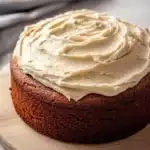 Banana bread birthday cake - the image is of a round cake on a wooden cutting board. the cake appears to be a chocolate cake with a golden brown crust. on top of the cake, there is a generous layer of creamy white frosting that is spread evenly across the cake. the frosting is smooth and smooth, and it looks like it has been freshly baked. the cutting board is placed on a gray tablecloth, and the background is blurred, making the cake the focal point of the image.