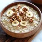 Banana bread oats - the image shows two bowls of oatmeal on a wooden cutting board. the oatmeal is a light brown color and appears to be thick and creamy. on top of the oatmeal, there are sliced bananas and walnuts arranged in a circular pattern. the bananas are cut into thin slices and are placed on top of each other. the walnuts are scattered around the bowl, adding a crunchy texture to the dish. the cutting board is placed on a white marble countertop.