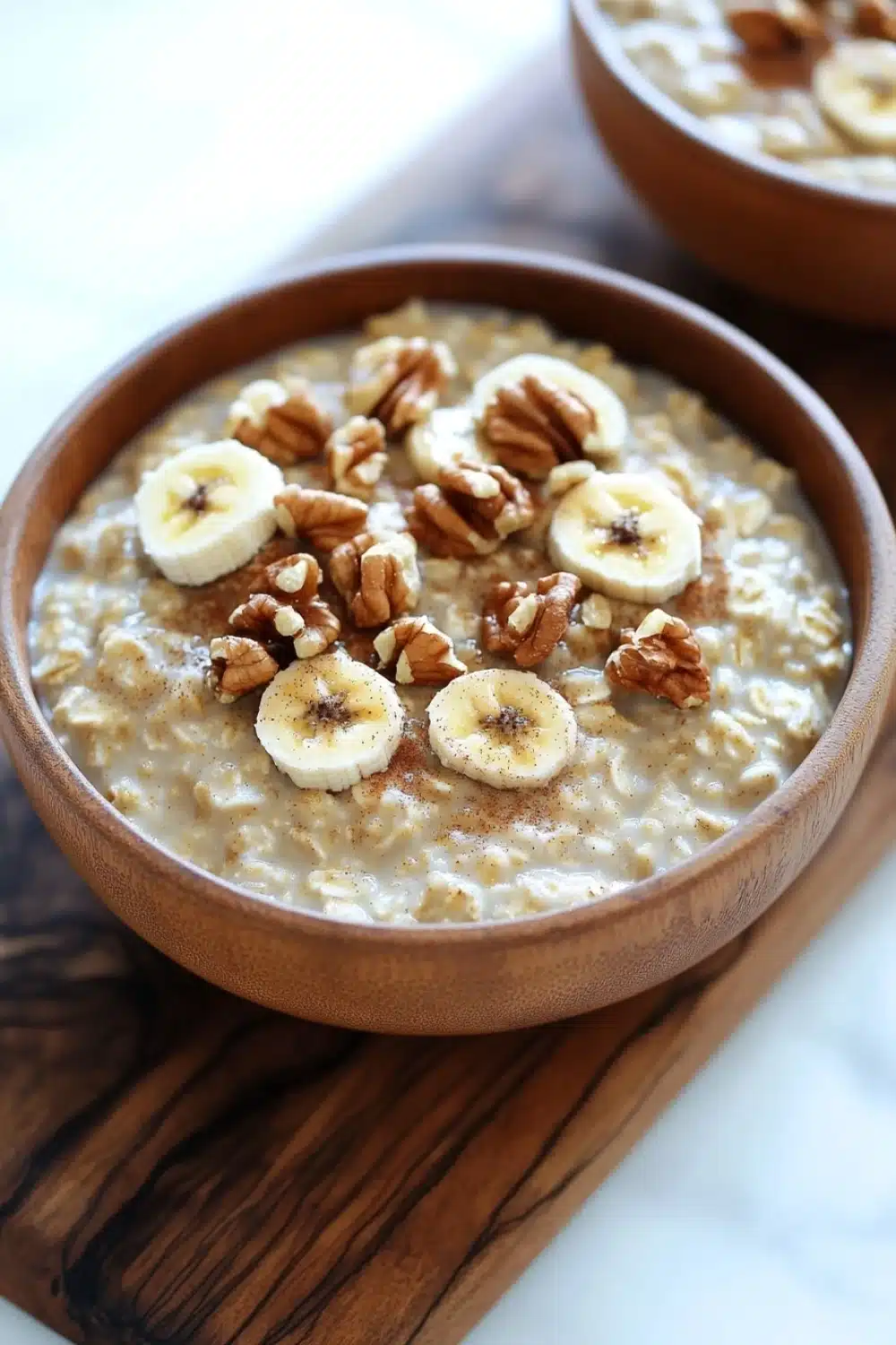 Banana bread oats - the image shows two bowls of oatmeal on a wooden cutting board. the oatmeal is a light brown color and appears to be thick and creamy. on top of the oatmeal, there are sliced bananas and walnuts arranged in a circular pattern. the bananas are cut into thin slices and are placed on top of each other. the walnuts are scattered around the bowl, adding a crunchy texture to the dish. the cutting board is placed on a white marble countertop.