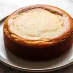 Banana cake allrecipes - the image is a close-up of a round cake on a white plate. the cake appears to be freshly baked and has a golden brown crust on top. the top of the cake is smooth and fluffy, with a light dusting of powdered sugar on top, giving it a golden-brown color. the plate is round and has thin edges. the background is a white marble countertop.