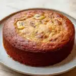 Banana cake made with cake mix - the image is of a round cake on a white plate. the cake appears to be freshly baked and has a golden brown color. it has a crumbly texture on top, with small pieces of chocolate chips scattered throughout. the plate is sitting on a wooden table with a light-colored surface. the background is blurred, making the cake the focal point of the image.
