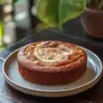 Banana cake snap - the image shows a round cake on a blue and white plate. the cake appears to be a bundt cake with a golden brown crust and a swirl of butter on top. the plate is placed on a wooden table with a potted plant in the background. the overall mood of the image is warm and inviting.