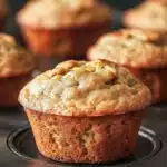 Banana muffin with applesauce and zucchini - the image shows a group of freshly baked muffins on a black baking tray. the muffins are golden brown in color and have a crumbly texture. one of the muffins has a bite taken out of it, revealing the soft, fluffy interior. the tray is lined with parchment paper and there are a few more muffins visible in the background. the overall mood of the image is warm and inviting.