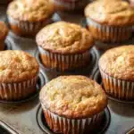 Banana muffin without egg - the image shows a tray of freshly baked muffins. the muffins are golden brown in color and have a crumbly texture on top. they are arranged in a single layer in the tray, with some overlapping each other. the tray is lined with parchment paper and is placed on a wooden table. the background is blurred, but it appears to be a kitchen countertop.