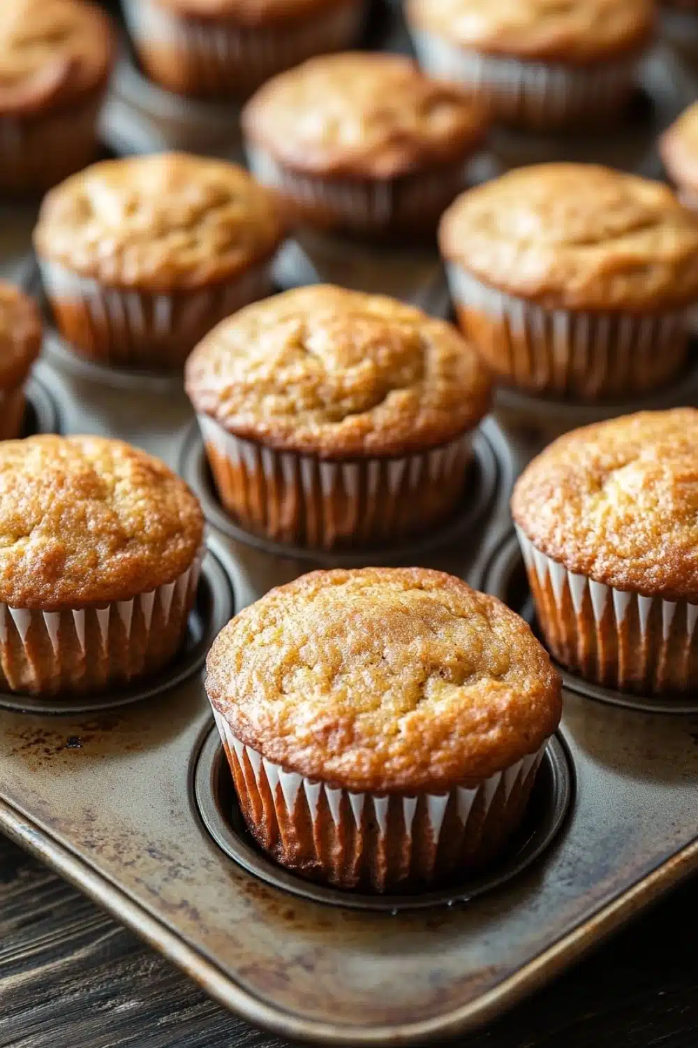 Banana muffin without egg - the image shows a tray of freshly baked muffins. the muffins are golden brown in color and have a crumbly texture on top. they are arranged in a single layer in the tray, with some overlapping each other. the tray is lined with parchment paper and is placed on a wooden table. the background is blurred, but it appears to be a kitchen countertop.