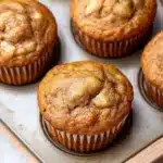 Banana muffin without egg - the image shows a muffin tin filled with freshly baked muffins. the muffins are golden brown in color and have a crumbly texture on top. they are arranged in a single layer in the tin, with some overlapping each other. the tin is placed on a wooden tray, and the background is blurred, but it appears to be a kitchen countertop.