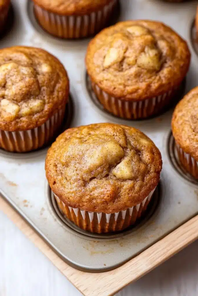 Banana muffin without egg - the image shows a muffin tin filled with freshly baked muffins. the muffins are golden brown in color and have a crumbly texture on top. they are arranged in a single layer in the tin, with some overlapping each other. the tin is placed on a wooden tray, and the background is blurred, but it appears to be a kitchen countertop.