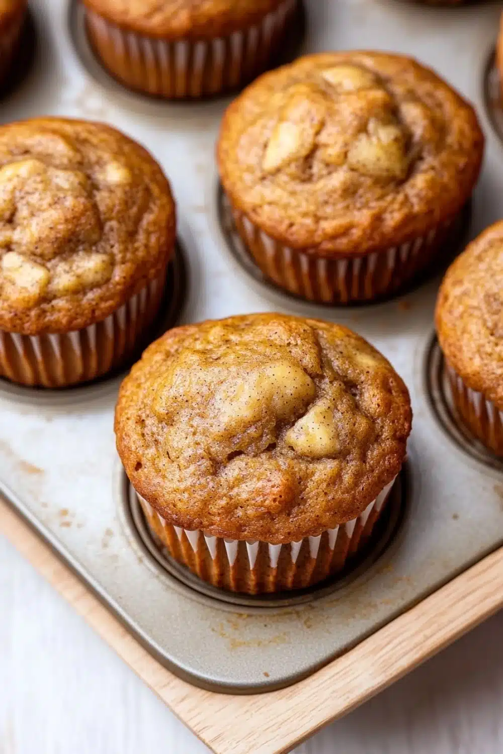 Banana muffin without egg - the image shows a muffin tin filled with freshly baked muffins. the muffins are golden brown in color and have a crumbly texture on top. they are arranged in a single layer in the tin, with some overlapping each other. the tin is placed on a wooden tray, and the background is blurred, but it appears to be a kitchen countertop.