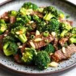Beef and broccoli stir fry spicy - the image is a close-up of a plate of stir-fry with beef and broccoli. the beef is cooked medium-rare and appears to be seasoned with sesame seeds and green onions. the broccoli is bright green and looks fresh and healthy. the plate is black with a speckled pattern and is sitting on a white marble countertop. the dish looks appetizing and ready to eat.