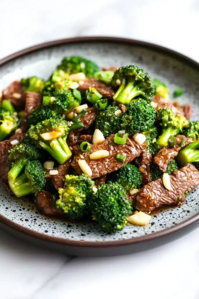 Beef and broccoli stir fry spicy - the image is a close-up of a plate of stir-fry with beef and broccoli. the beef is cooked medium-rare and appears to be seasoned with sesame seeds and green onions. the broccoli is bright green and looks fresh and healthy. the plate is black with a speckled pattern and is sitting on a white marble countertop. the dish looks appetizing and ready to eat.