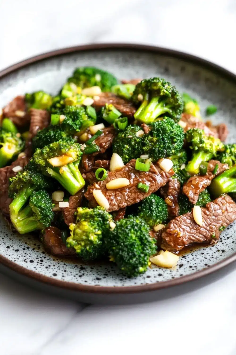 Beef and broccoli stir fry spicy - the image is a close-up of a plate of stir-fry with beef and broccoli. the beef is cooked medium-rare and appears to be seasoned with sesame seeds and green onions. the broccoli is bright green and looks fresh and healthy. the plate is black with a speckled pattern and is sitting on a white marble countertop. the dish looks appetizing and ready to eat.