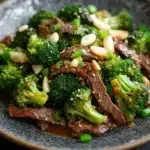 Beef and broccoli stir fry spicy - the image is a close-up of a plate of stir-fry with beef and broccoli. the beef is cooked medium-rare and appears to be seasoned with sesame seeds and sliced almonds. the broccoli is bright green and looks fresh and healthy. the plate is black and has a textured surface. the background is blurred, making the plate the focal point of the image.