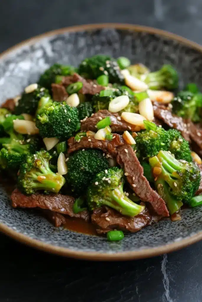 Beef and broccoli stir fry spicy - the image is a close-up of a plate of stir-fry with beef and broccoli. the beef is cooked medium-rare and appears to be seasoned with sesame seeds and sliced almonds. the broccoli is bright green and looks fresh and healthy. the plate is black and has a textured surface. the background is blurred, making the plate the focal point of the image.