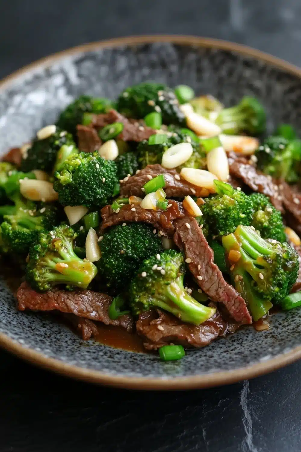 Beef and broccoli stir fry spicy - the image is a close-up of a plate of stir-fry with beef and broccoli. the beef is cooked medium-rare and appears to be seasoned with sesame seeds and sliced almonds. the broccoli is bright green and looks fresh and healthy. the plate is black and has a textured surface. the background is blurred, making the plate the focal point of the image.