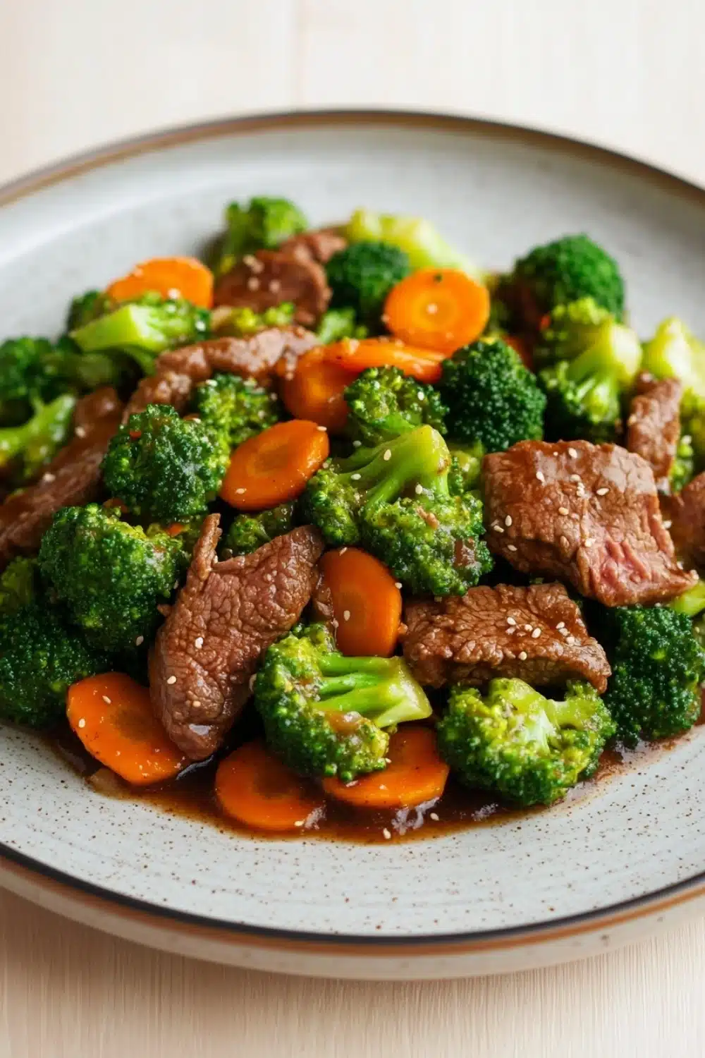 Beef and broccoli stir fry with carrots - the image is a close-up of a plate of stir-fry with beef and broccoli. the beef is cooked medium-rare and appears to be seasoned with herbs and spices. the broccoli is bright green and looks fresh and healthy. there are also small pieces of orange carrots scattered throughout the dish. the plate is white with a speckled pattern and is sitting on a wooden table.