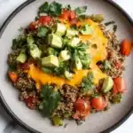 Beef quinoa taco skillet - the image is a close-up of a plate of food. the plate is round and has a gray rim. the food appears to be a quinoa salad with diced tomatoes, avocado, and cilantro scattered throughout. the quinoa is a light brown color and the avocado is cut into small cubes. the avocado is a bright yellow color and is placed on top of the quinoa. the cilantro is a dark green color and adds a pop of color to the dish. the background is a white marble countertop.