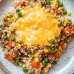 Beef rice veggie casserole - the image is a close-up of a plate of food. the plate is round and has a light blue rim with a speckled pattern. the food appears to be a mixture of rice, ground beef, peas, carrots, corn, and other vegetables. on top of the rice, there is a layer of melted cheese. the cheese is a bright yellow color and is spread evenly over the top. the colors of the food are vibrant and the dish looks appetizing.