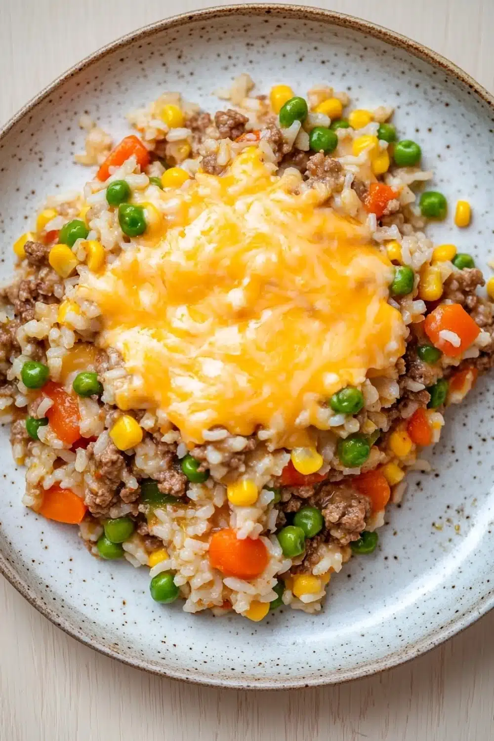 Beef rice veggie casserole - the image is a close-up of a plate of food. the plate is round and has a light blue rim with a speckled pattern. the food appears to be a mixture of rice, ground beef, peas, carrots, corn, and other vegetables. on top of the rice, there is a layer of melted cheese. the cheese is a bright yellow color and is spread evenly over the top. the colors of the food are vibrant and the dish looks appetizing.