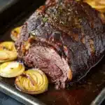 Beef roast crockpot slow cooker au jus - the image shows a large piece of roast beef on a baking tray. the roast beef is cooked medium-rare and has a dark brown glaze on top. it is garnished with fresh herbs and is surrounded by sliced onions. the tray is lined with parchment paper and is placed on a dark grey countertop. the overall presentation of the dish is elegant and appetizing.