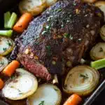Beef sirloin tip roast slow cooker - the image shows a large piece of cooked meat on a black baking tray. the meat appears to be a roast beef, with a dark brown crust on top and a juicy center. it is garnished with chopped herbs and spices, and is surrounded by sliced carrots, celery, and onions. the vegetables are arranged in a circular pattern around the meat, with some overlapping each other. the tray is sitting on a wooden table, and the background is blurred.