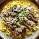 Beef stroganoff crockpot freezer meal - the image shows a plate of beef stroganoff with fettuccine noodles. the dish is served in a brown ceramic bowl with a white rim. the beef is cooked medium-rare and is covered in a creamy mushroom sauce. the sauce is drizzled over the top of the beef and is garnished with chopped parsley. the noodles are long and thin, and they are arranged in a pile on top. the plate is sitting on a wooden table.