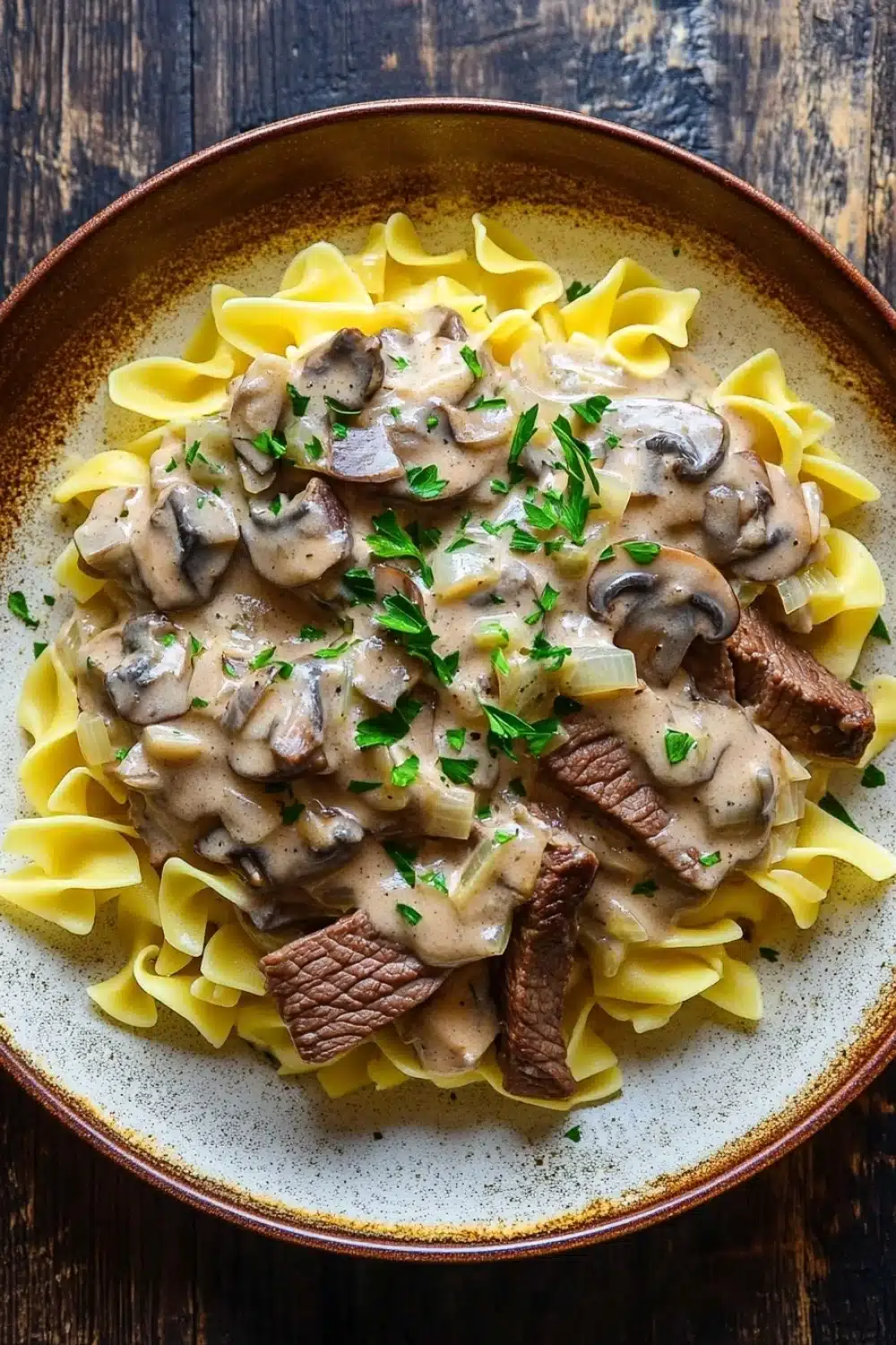 Beef stroganoff crockpot freezer meal - the image shows a plate of beef stroganoff with fettuccine noodles. the dish is served in a brown ceramic bowl with a white rim. the beef is cooked medium-rare and is covered in a creamy mushroom sauce. the sauce is drizzled over the top of the beef and is garnished with chopped parsley. the noodles are long and thin, and they are arranged in a pile on top. the plate is sitting on a wooden table.