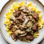 Beef stroganoff crockpot freezer meal - the image shows a plate of beef stroganoff with fettuccine noodles. the beef is cooked medium-rare and is covered in a creamy mushroom sauce. the noodles are long and thin, and they are arranged in a pile on top of the beef. the dish is garnished with chopped parsley and sliced onions. the plate is sitting on a white marble countertop.