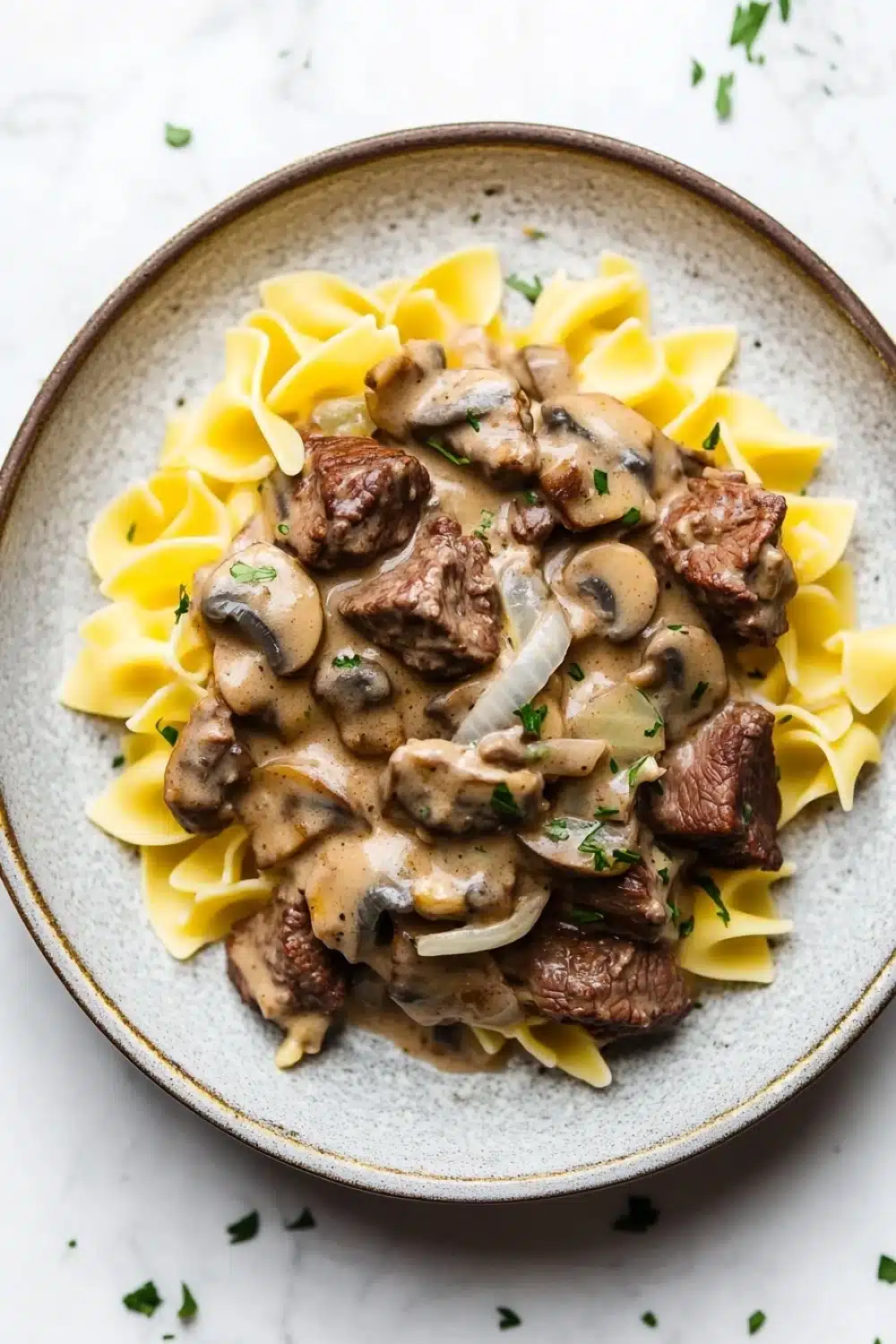 Beef stroganoff crockpot freezer meal - the image shows a plate of beef stroganoff with fettuccine noodles. the beef is cooked medium-rare and is covered in a creamy mushroom sauce. the noodles are long and thin, and they are arranged in a pile on top of the beef. the dish is garnished with chopped parsley and sliced onions. the plate is sitting on a white marble countertop.