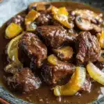 Beef tips freezer meal - the image is a close-up of a dish in a bowl. the dish appears to be a beef stew with chunks of beef, mushrooms, and yellow bell peppers. the beef is cooked in a rich, dark brown sauce and is garnished with sliced onions and mushrooms. the bowl is sitting on a wooden table with a dark background. the overall color scheme of the dish is warm and inviting.