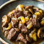 Beef tips freezer meal - the image shows a plate of beef stew with mushrooms and onions. the beef appears to be cooked medium-rare and is covered in a rich, dark brown sauce. the mushrooms are sliced and arranged in a circular pattern around the beef. the onions are bright yellow and appear to be caramelized. the dish is served on a dark brown plate with a gold rim. the background is blurred, but it seems to be a wooden table.