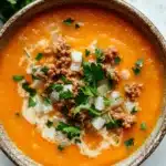 Butternut squash soup ground turkey - the image is a close-up of a bowl of tomato soup. the soup is a bright orange color and appears to be thick and creamy. it is topped with chunks of ground meat, diced onions, and chopped parsley. the bowl is made of ceramic and is sitting on a white marble countertop. there are a few sprigs of fresh parsley scattered around the bowl.