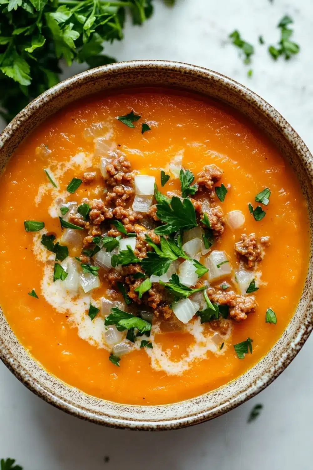 Butternut squash soup ground turkey - the image is a close-up of a bowl of tomato soup. the soup is a bright orange color and appears to be thick and creamy. it is topped with chunks of ground meat, diced onions, and chopped parsley. the bowl is made of ceramic and is sitting on a white marble countertop. there are a few sprigs of fresh parsley scattered around the bowl.