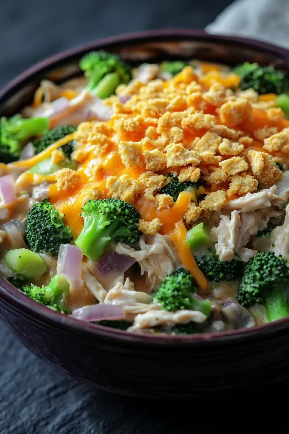Chicken broccoli casserole mushroom soup - the image is a close-up of a bowl of food. the bowl is made of dark brown ceramic and is sitting on a dark grey countertop. the food appears to be a stir-fry with broccoli, chicken, and other vegetables mixed in. the broccoli is bright green and looks fresh, while the chicken is white and fluffy. the chicken is cut into small pieces and is scattered throughout the dish. on top of the chicken, there is a layer of melted cheese, which is a bright orange color. the cheese is crumbled and golden brown, and it looks like it has been cooked until it is melted and bubbly. there are also some small pieces of red onions and green onions scattered throughout. the dish looks appetizing and ready to eat.