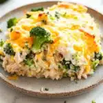 Chicken broccoli rice casserole greek yogurt - the image shows a plate of food with a serving of rice and broccoli. the rice appears to be cooked and has a golden brown color. on top of the rice, there is a layer of melted cheese, which is a bright yellow color. the broccoli is cut into small pieces and is scattered around the plate. the plate is on a white marble countertop with a few pieces of broccoli scattered around. the background is blurred, but it seems to be a kitchen countertop.