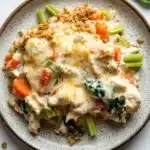 Chicken casserole greek yogurt - the image shows a plate of food on a white marble countertop. the plate is round and has a light brown speckled pattern. on top of the plate, there is a serving of chicken casserole with a golden brown crumbly topping. the chicken is covered in a creamy white sauce and is garnished with sliced carrots, celery, and spinach. there are also some fresh spinach leaves scattered around the plate. the dish appears to be freshly cooked and ready to eat.