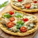 Chicken pesto pizza on naan bread - the image shows two small pizzas on a wooden cutting board. the pizzas are topped with shredded chicken, cherry tomatoes, and fresh basil leaves. the chicken is cooked to a golden brown color and appears to be seasoned with herbs and spices. the cherry tomatoes are bright red and look ripe and juicy. the basil leaves are scattered around the pizzas, adding a pop of green color to the dish. the cutting board is placed on a white marble countertop.