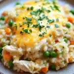Chicken rice casserole instant pot - the image shows a plate of risotto with chicken and peas. the risotto is a creamy yellow color and appears to be cooked until it is soft and fluffy. the chicken is cut into small pieces and is covered in a layer of melted cheese. the peas are bright green and are scattered throughout the risotto. the dish is garnished with a sprig of fresh parsley. the plate is sitting on a wooden table.