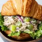 Chicken salad sandwich on croissant - the image shows a close-up of a croissant sandwich in a wooden bowl. the sandwich is made with a toasted bun and is filled with a variety of ingredients. the main focus of the image is on the sandwich, which appears to be made with chicken, lettuce, red onion, and green olives. the chicken is cut into small pieces and is mixed with a creamy dressing. the lettuce is a mix of green and red, and the olives are a bright green color. the bowl is sitting on a white marble countertop.