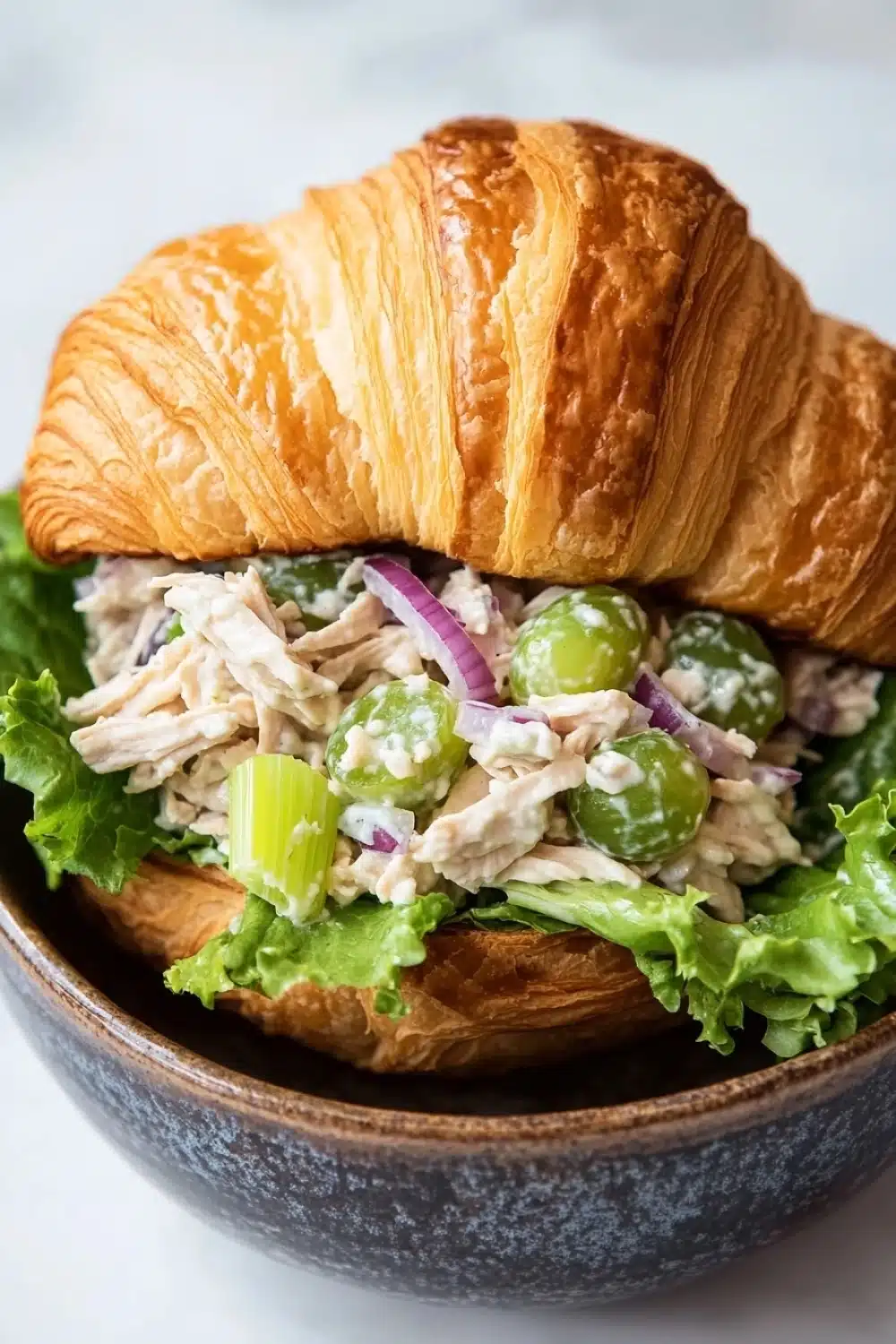 Chicken salad sandwich on croissant - the image shows a close-up of a croissant sandwich in a wooden bowl. the sandwich is made with a toasted bun and is filled with a variety of ingredients. the main focus of the image is on the sandwich, which appears to be made with chicken, lettuce, red onion, and green olives. the chicken is cut into small pieces and is mixed with a creamy dressing. the lettuce is a mix of green and red, and the olives are a bright green color. the bowl is sitting on a white marble countertop.