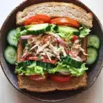 Chicken salad sandwich with veggies - the image shows a sandwich in a brown ceramic bowl. the sandwich is made with two slices of toasted bread and is filled with a variety of ingredients. on top of the bread, there are shredded chicken, sliced cucumbers, red onions, and lettuce. the vegetables are arranged in a colorful and appetizing manner. the bowl is sitting on a white wooden table.
